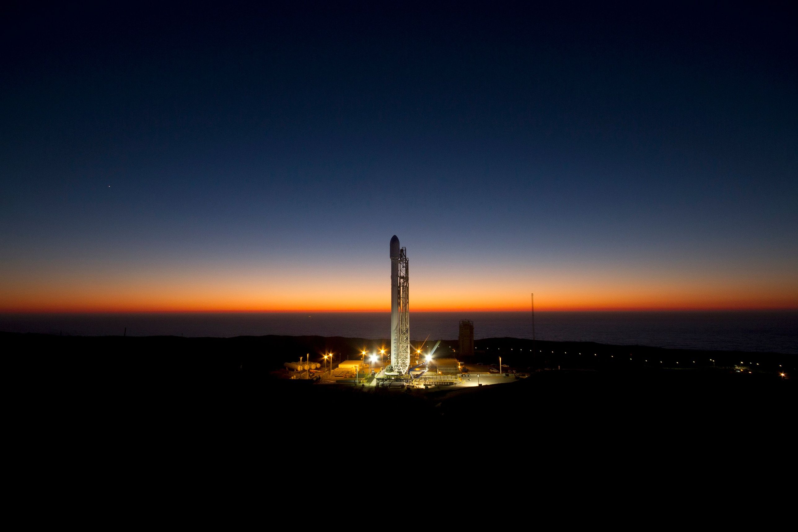 A rocket on its launch pad at dusk, lit against a dark blue sky over the sea
